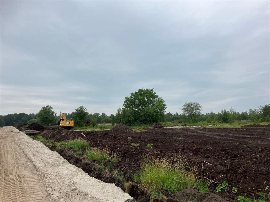 Graafmachine werkt aan het herstel van veengrond in het Bargerveen. Op de foto zie je omgewoelde zwarte grond, een zandpad en bomen op de achtergrond.