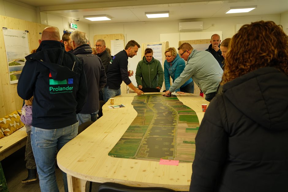 Bezoekers buigen zich over een houten tafel met een luchtfoto van het gebied. Een medewerker van Prolander wijst iets aan.