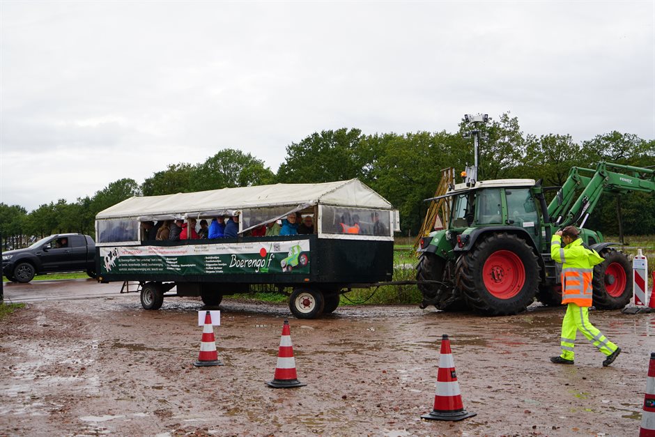 Bezoekers zitten in een huifkar achter een trekker op een modderig werkterrein. Een verkeersregelaar staat erbij.